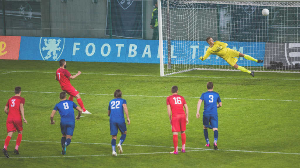 Soccer players watching as one player shoots a ball past a diving goalie